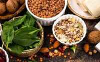Assortment of plant-based protein foods including lentils, spinach, seeds, nuts and tofu in bowls on a dark surface.