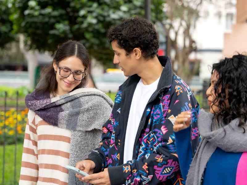 Three students standing together outside, talking and checking a phone. latin america