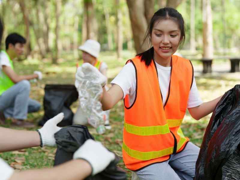 Young volunteer wearing a reflective vest picking up plastic waste in a park during a cleanup activity.