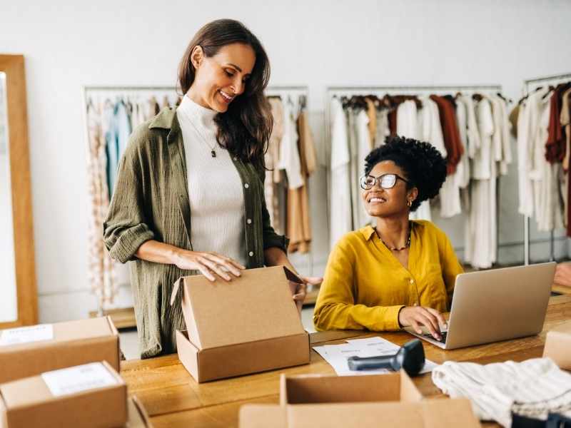 Two women at a workspace preparing packages and organizing online orders in a boutique setting.