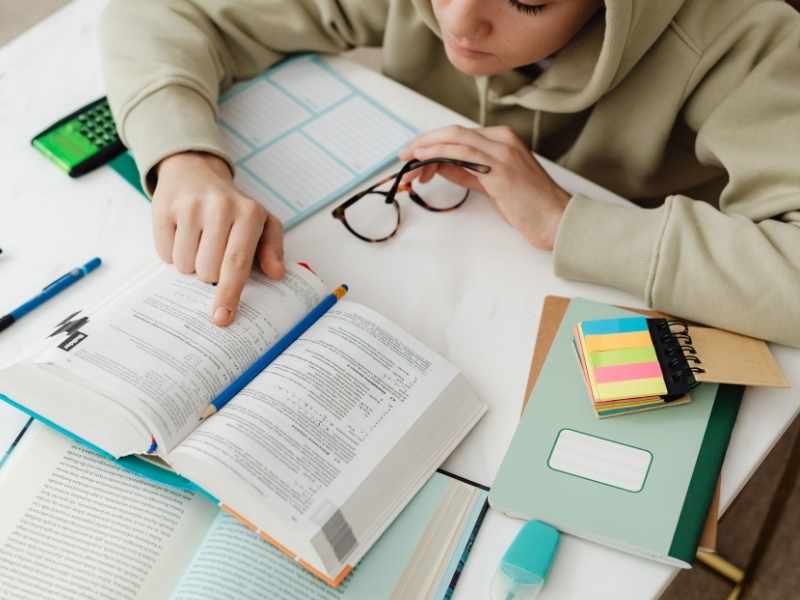 Person studying an open book with sticky notes, highlighters, and reading glasses nearby.