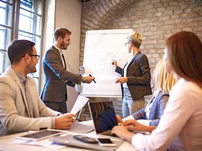 Team in a meeting room with laptops while a presenter speaks next to a flip chart during a business discussion.